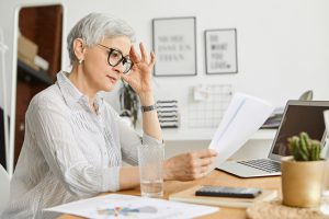 People, aging, technology and profession concept. Serious 50 year old Caucasian female wearing stylish eyeglasses and silk shirt reading contract while working at desk, sitting in front of open laptop | Pension de reversion attestation fiscale pension de réversion
