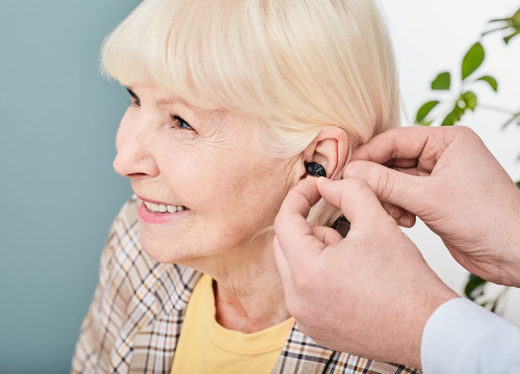 Audiologist inserting Intra-ear hearing aid to senior woman patient, close-up. Hearing solution for elderly people | Pension de reversion femme senior handicapée prothèse auditive
