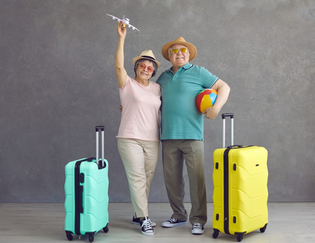 Studio portrait of happy senior couple in hats and sunglasses with paper plane and suitcases | Pension de reversion Seniors avec valises partant en vacances