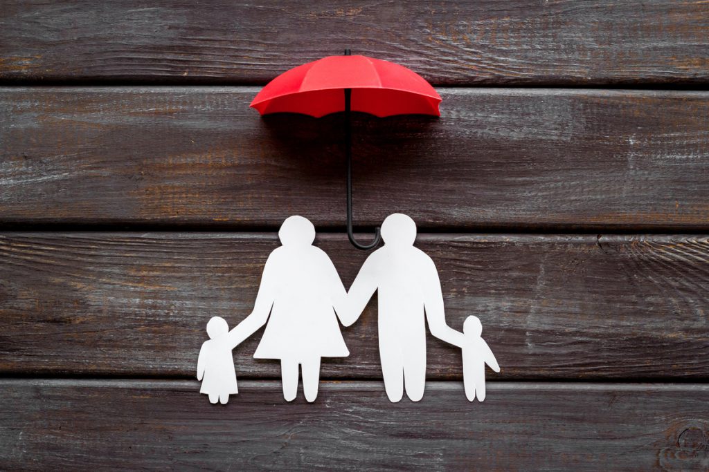 Health insurance concept. Family cutout under umbrella on dark wooden background top-down copy space | Pension de reversion capital décès Ircantec