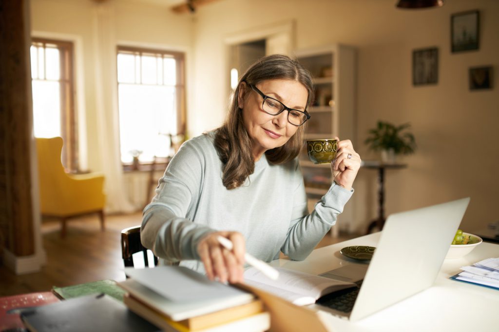 Serious confident middle aged female journalist with cup of coffee sitting at desk with books and laptop, making research, studying literature. Attractive mature woman copywriter working from home | Pension de reversion document perdu demande de retraite