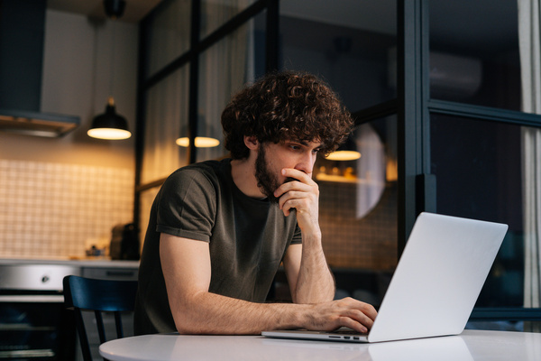 Side view of thoughtful young man programming on laptop sitting at table in kitchen room with modern interior. | Pension de reversion