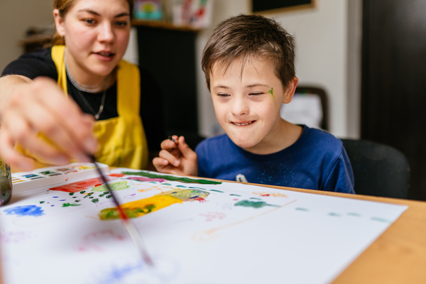 Young female teacher explain, talk at desk with a down syndrome schoolboy. Color painting for disabled kids, child who are down syndrome and student teacher, development of children with spesial needs | Pension de reversion trimestres éducation enfant handicapé