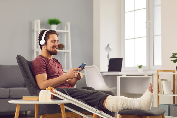 Young man with broken leg sitting in chair and listening to music on mobile phone | Pension de reversion droits retraite arrêt maladie