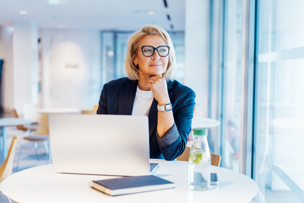 50's confident mature businesswoman dreamy looking at window, middle-aged experienced senior female professional working on laptop in open space office. Female entrepreneur working remotely. | Pension de reversion corriger son relevé de carrière