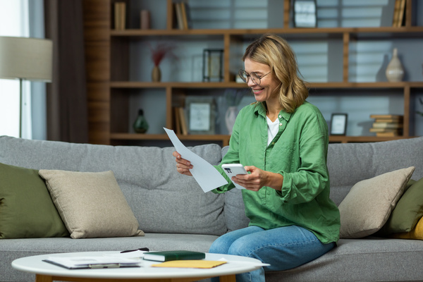 An elderly woman received good news, financial deal, loan repayment, house sale, pension, subsidy. He is sitting on the sofa at home, holding the phone and documents in his hands. Checks accounts | Pension de reversion petite pension augmentée