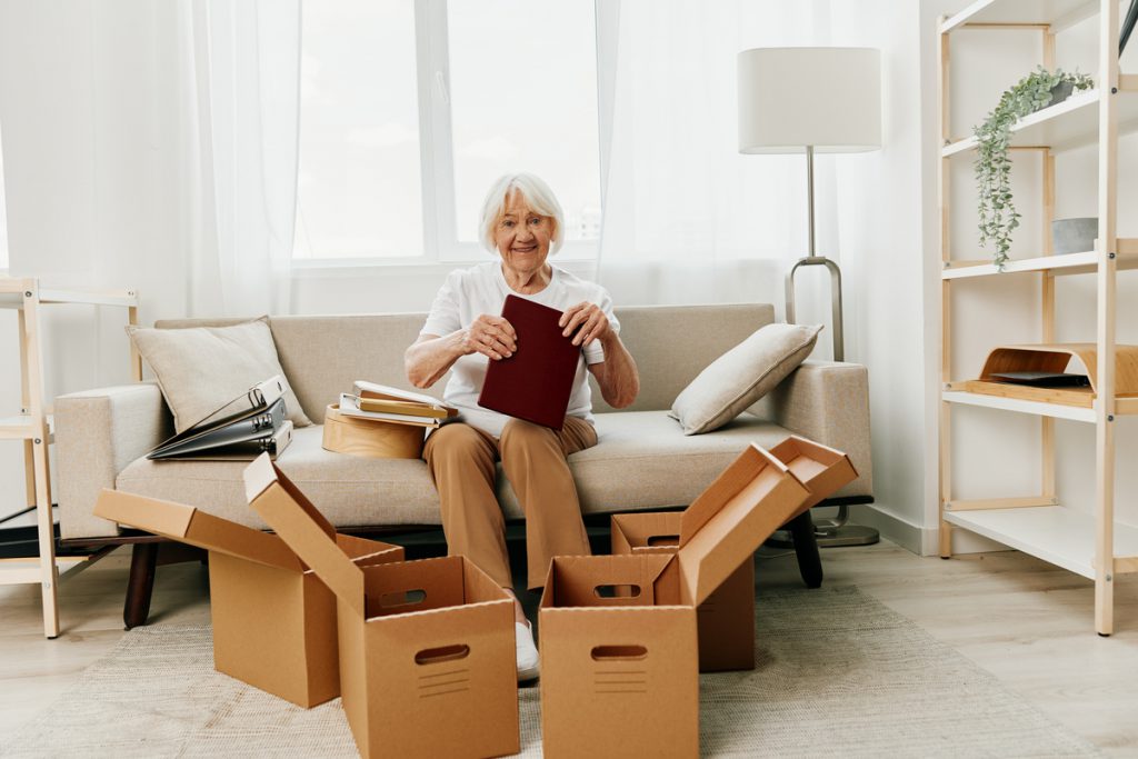 elderly woman sits on a sofa at home with boxes. collecting things with memories and moving and happiness smile. | Pension de reversion résidence autonomie