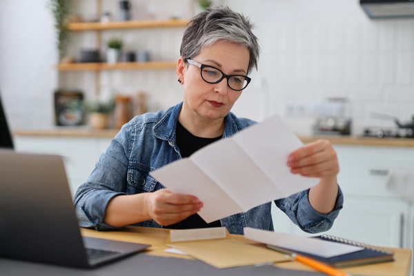 Serious mature woman in glasses reading the bill in the kitchen. | Pension de reversion attestation fiscale retraite perdue