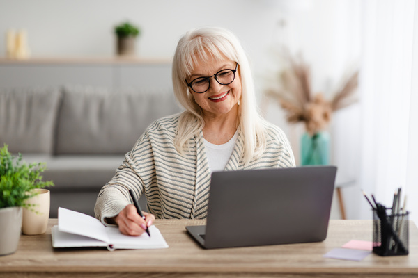Mature woman in glasses writing and using laptop at home | Pension de reversion calendrier paiement retraite fonction publique