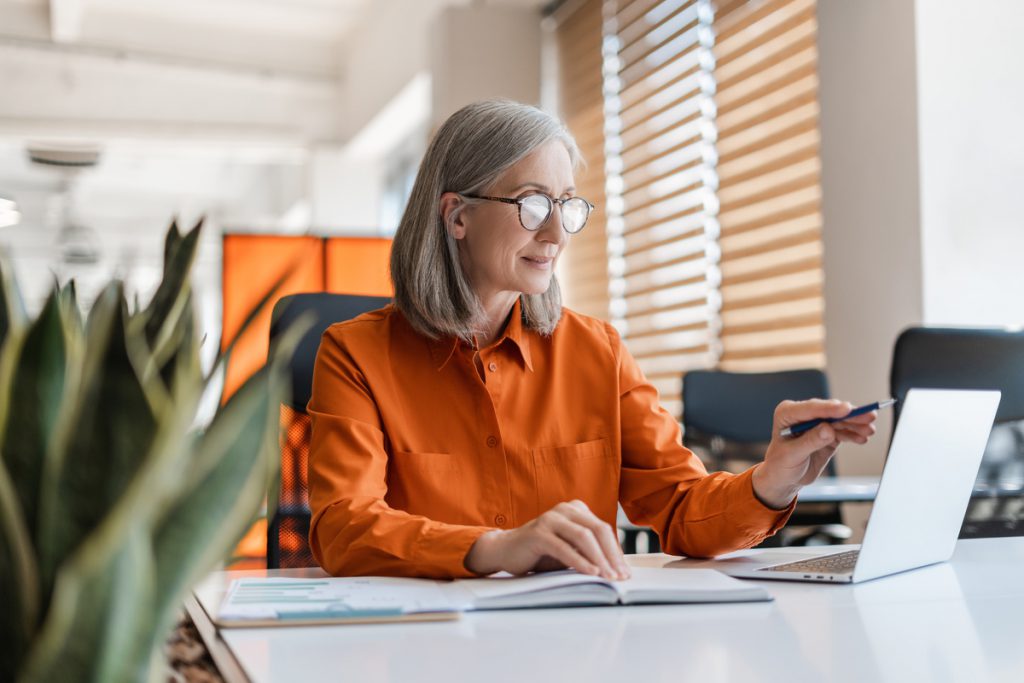 Portrait of beautiful confident senior woman, manager using laptop computer working online sitting in modern office. Business woman wearing stylish eyeglasses checking email. Technology concept | Pension de reversion espace personnel Agirc-Arrco