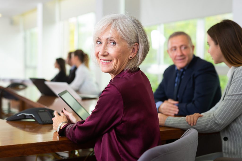 Smiling mature senior white businesswoman posing sitting at table | Pension de reversion prime emploi seniors