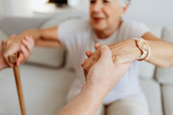Cropped shot a young male home carer supporting old woman to stand up from the sofa at care home. Photo of male professional caregiver taking care of elderly woman at home during the day. | Pension de reversion dispense de frais d'hébergement