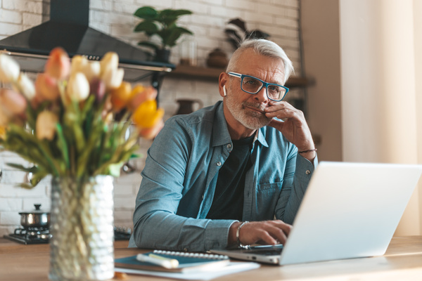 A mature entrepreneur in a home interior works remotely on a laptop. Portrait of a senior man with a beard wearing glasses. | Pension de reversion refus de retraite progressive