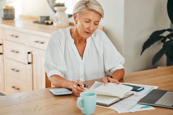 Schedule, documents and finance with a senior woman planning for her future retirement with savings, investment and wealth. Notebook, writing and laptop with an elderly female pensioner at home | Pension de reversion retraite et trop-perçu France Travail