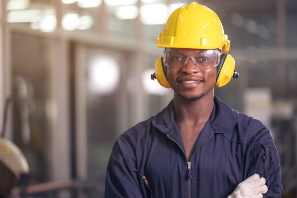 Portrait of cheerful black worker wearing protective headphones posing looking at camera and enjoying work at background factory | Pension de reversion retraite anticipée grâce aux points de pénibilité