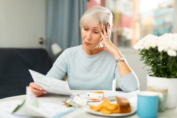 Upset senior woman looking worriedly at papers at home table | Pension de reversion retard paiement retraite complémentaire