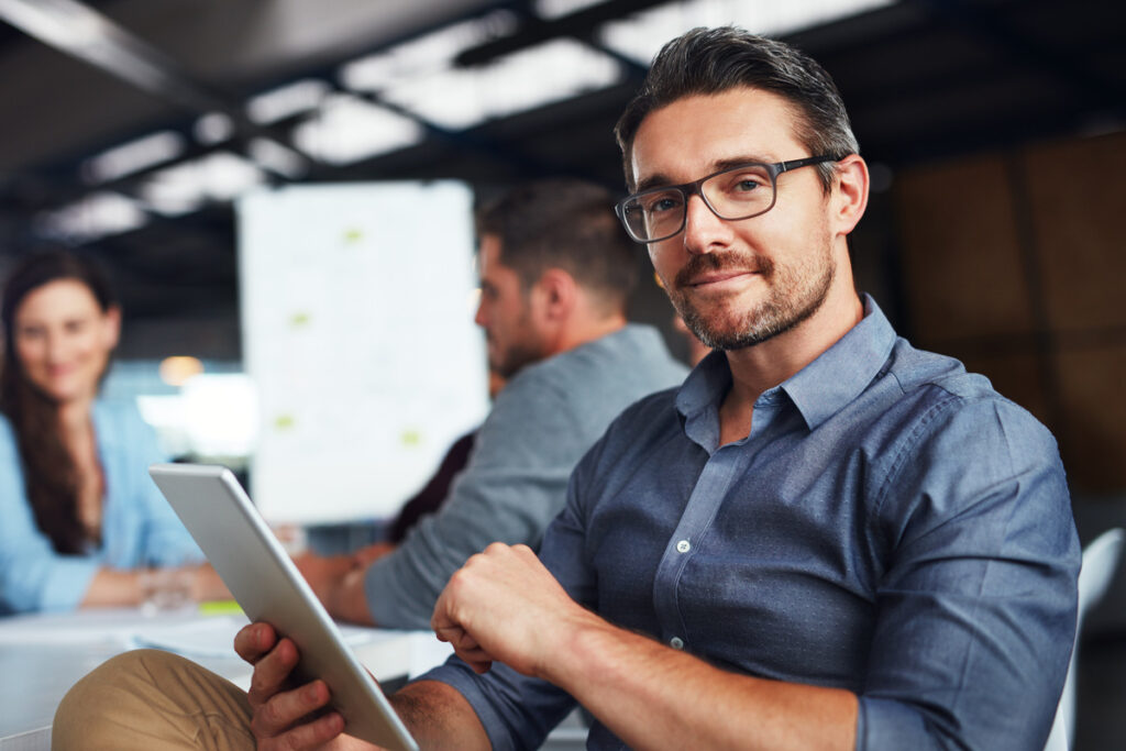 The creative process Portrait of a man sitting at a table in an office using a digital tablet with colleagues working in the background | Pension de reversion validation trimestre retraite