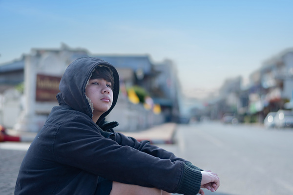 Asian young boy wearing a dark hooded jacket sitting near a stone fence or barrier thoughtfully with late morning road background emphasizing introspective unhappy sad mood concept | Pension de reversion pension de réversion orphelin