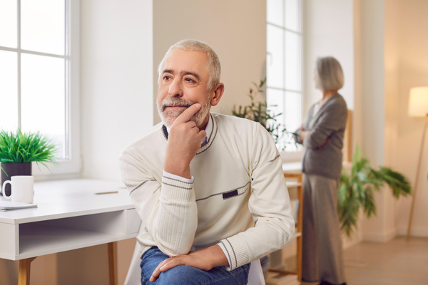 Upset senior old man sitting on chair with a woman wife in background ignoring each other at home | Pension de reversion divorce retraite