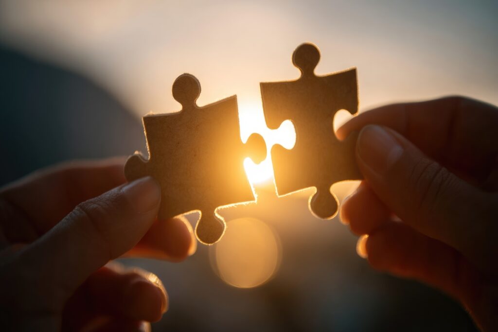 Two hands hold puzzle pieces against glowing sunset Symbol of connection harmony Fitting together life Teamwork relationships problem-solving Team work concept Sun light sky | Pension de reversion Symbole de la pension de réversion Agirc-Arrco versée au conjoint survivant.