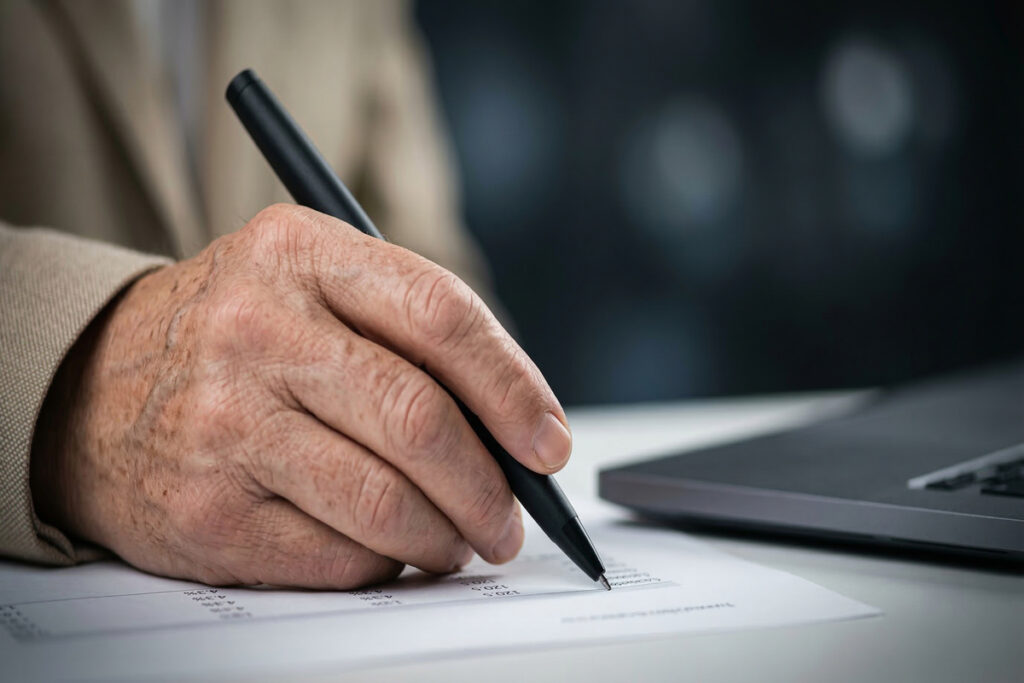 Senior man hand signing a document with a black pen | Pension de reversion Senior vérifiant ses comptes et remplissant un formulaire officiel pour une réclamation de pension.