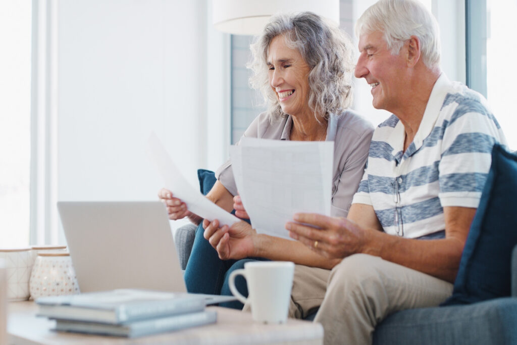 Laptop document and old couple in house with smile life insurance website or retirement planning Married senior people and reading paperwork on sofa with computer estate policy and pension fund | Pension de reversion Simulation sur Info-Retraite pour sécuriser ses droit à la majoration de pension de réversion.