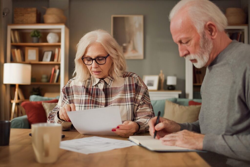 Seniors review life insurance options while sitting at wooden table in cozy living room filled with sunlight | Pension de reversion Couple de retraités analysant des documents financiers sur le rachat de trimestres et la réversion