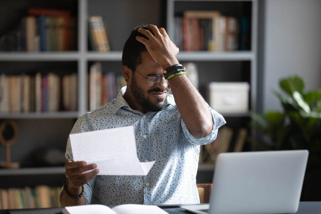 Stressed biracial man frustrated by bad news in letter | Pension de reversion Un homme inquiet dans son bureau lit un courrier de notification de trop-perçu après une erreur administrative.