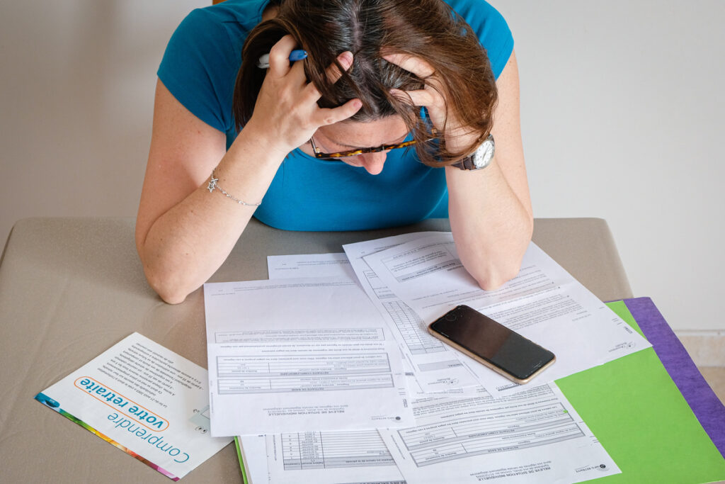 Retraite-administration-calculer | Pension de reversion Une personne en détresse, tête baissée et mains sur le visage, assise à une table croulant sous des documents officiels, un smartphone et des dossiers de l'Assurance Retraite. Illustration de la difficulté à faire valoir ses droits à la réversion militaire.
