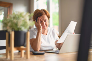 Femme âgée découragée face à des documents administratifs pour la régularisation de la réversion. | Pension de reversion Une femme âgée assise à une table, la main sur le front, désemparée, tenant une lettre en main avec un ordinateur et une calculatrice devant elle, illustrant le besoin de régularisation de la réversion.